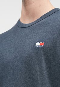 Detailní záběr na osobu v modrém tričku Tommy Jeans s malým logem v červené, bílé a námořnické modři na hrudi.