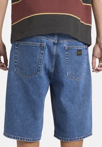 Rückansicht einer Person, die blaue Jeansshorts mit zwei Gesäßtaschen trägt, von denen eine mit einem kleinen schwarzen und gelben RVCA-Logo-Patch versehen ist.
