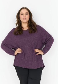 Lila, hosszú ujjú felső V-kivágással, laza szab�ással, texturált anyagból, lekerekített hemél. Fekete nadrággal párosítva.