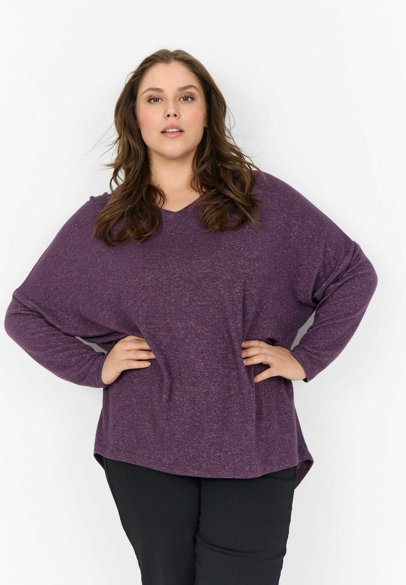 Lila, hosszú ujjú felső V-kivágással, laza szabással, texturált anyagból, lekerekített hemél. Fekete nadrággal párosítva.