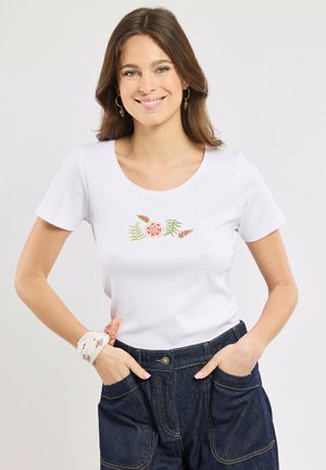 Femme portant une chemise blanche à manches courtes avec une broderie florale rose, un pantalon en denim foncé, des boucles d'oreilles créoles dorées et un foulard de poignet blanc à motifs.