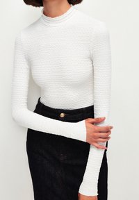 Top bianco a maniche lunghe con tessuto texturizzato e ondulato, con colletto falso. Abbinato a una gonna in denim scuro con dettaglio di bottoni frontali.