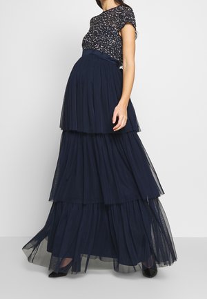 Donna che indossa una gonna lunga a balze blu navy e una maglietta nera a maniche corte con paillettes, in piedi davanti a uno sfondo chiaro uniforme.