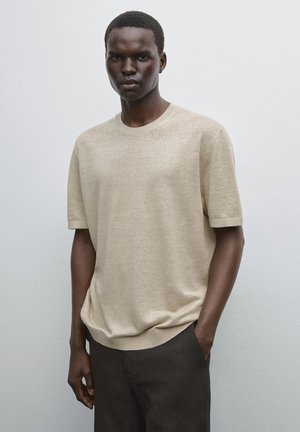 Beige Strick-T-Shirt mit kurzen Ärmeln, Rundhalsausschnitt und gerippten Akzenten, kombiniert mit dunkelbraunen Hosen. Glatte Textur, lockere Passform.