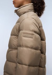 Beige pufferjack met een hoge kraag, voorzien van doorgestikte secties en een merkembleem op de mouw. Glad, glanzend textuur.