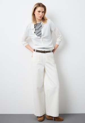 Jeune femme aux cheveux blonds posant devant un mur blanc, portant une chemise blanche ouverte, un haut gris, un pantalon blanc à jambes larges, une ceinture marron et des chaussures marron.