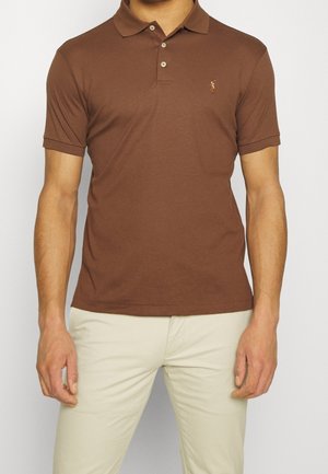 Mand iført brun kortærmet polo med tre knapper og beige bukser, stående mod en ensfarvet lys baggrund.