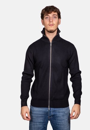 Maglione a costine neri con zip alta, dotato di una zip argento, maniche lunghe e un design aderente. Indossato sopra a jeans blu.