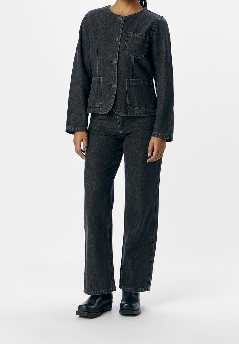 Personne portant une veste en jean noire avec des boutons sur le devant, trois poches plaquées, un jean droit assorti et des chaussures à plateforme noires.