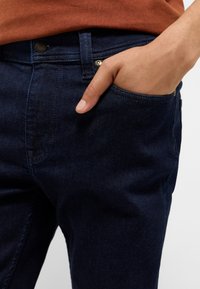 Granatowe dżinsy z prostym krojem, z przednimi kieszeniami i metalowym zapięciem na guzik w pasie. Gładka tekstura.