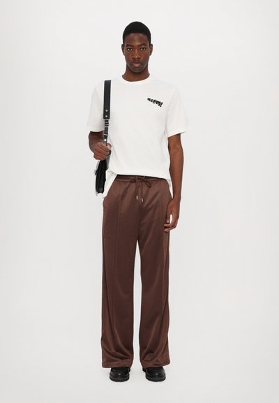 Weißes T-Shirt mit schwarzem Grafikdesign, braune Jogginghose mit Kordelzug und schwarze Stiefel. Hält eine schwarze Umhängetasche. Schlichtes, lässiges Design.