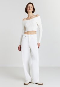 Weißer gerippter, schulterfreier, kurzes Sweatshirt, kombiniert mit hochtaillierten, weißen weit geschnittenen Jeans und crèmefarbigen Sneakers mit braunen Akzenten.