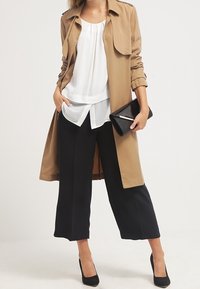 Beiger Trenchcoat über einer weißen Bluse mit Bindegürtel, schwarze 7/8-Hosen und schwarze High Heels, hält eine schwarze Clutch.