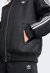 Une personne portant une veste bomber noire avec une fermeture éclair, ornée de rayures blanches diagonales et d'un logo Adidas, les mains dans les poches.