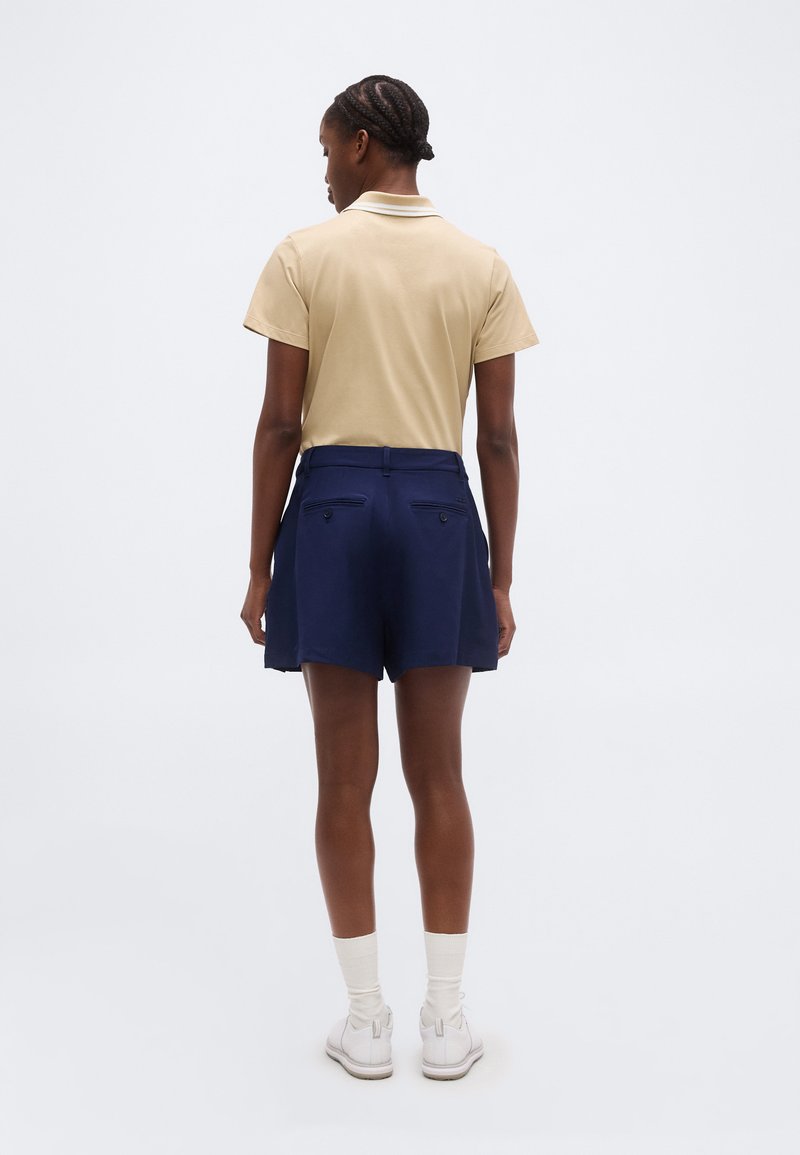 Femme aux cheveux tressés portant un polo beige à manches courtes, un short taille haute bleu marine, des chaussettes blanches et des baskets blanches, debout de dos.