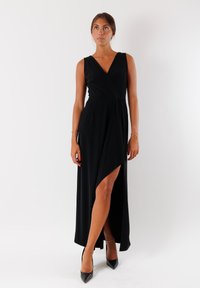 Abito nero senza maniche con un design avvolgente, scollatura a V profonda, alta spacco frontale e texture liscia. Abbinato a décolleté nere con punta appuntita.