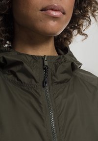 Olivgrön zip-up jacka med hög krage, glänsande dragkedja och en lätt, slät textur. Huva fastsydd, minimalistisk design.