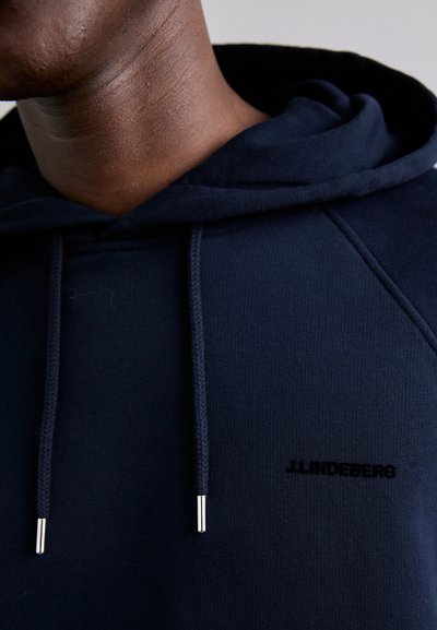 Felpa blu navy con una texture morbida, dotata di cordini con punte in argento e un piccolo logo nero. Polsini e orlo elastici.