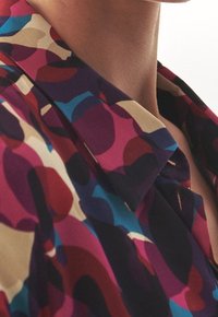 Blusa multicolore con un audace motivo astratto in tonalità di viola, rosso, blu e beige, dotata di un collo classico e una morbida texture del tessuto.