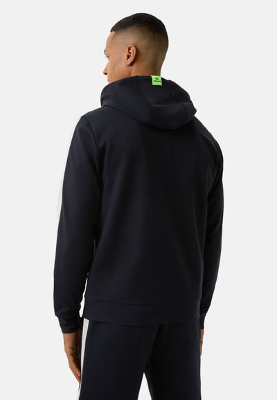 Marineblaue Kapuzenjacke mit lockerer Passform, versehen mit einem neongrünen Etikett auf der Rückseite und strukturiertem Stoff. Lange Ärmel mit elastischen Bündchen.