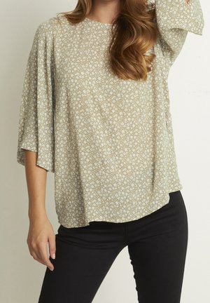 Bluse mit Blumenmuster in Hellgrün und Beige, mit Glockenärmeln und rundem Halsausschnitt, kombiniert mit schwarzen, figurbetonten Hosen.