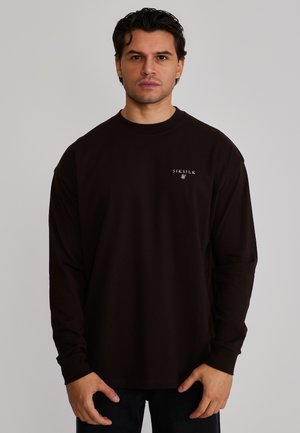 Mand med mørkt hår iført en sort langærmet skjorte med "SIKSILK"-logo på brystet, stående mod en ensfarvet lys baggrund.