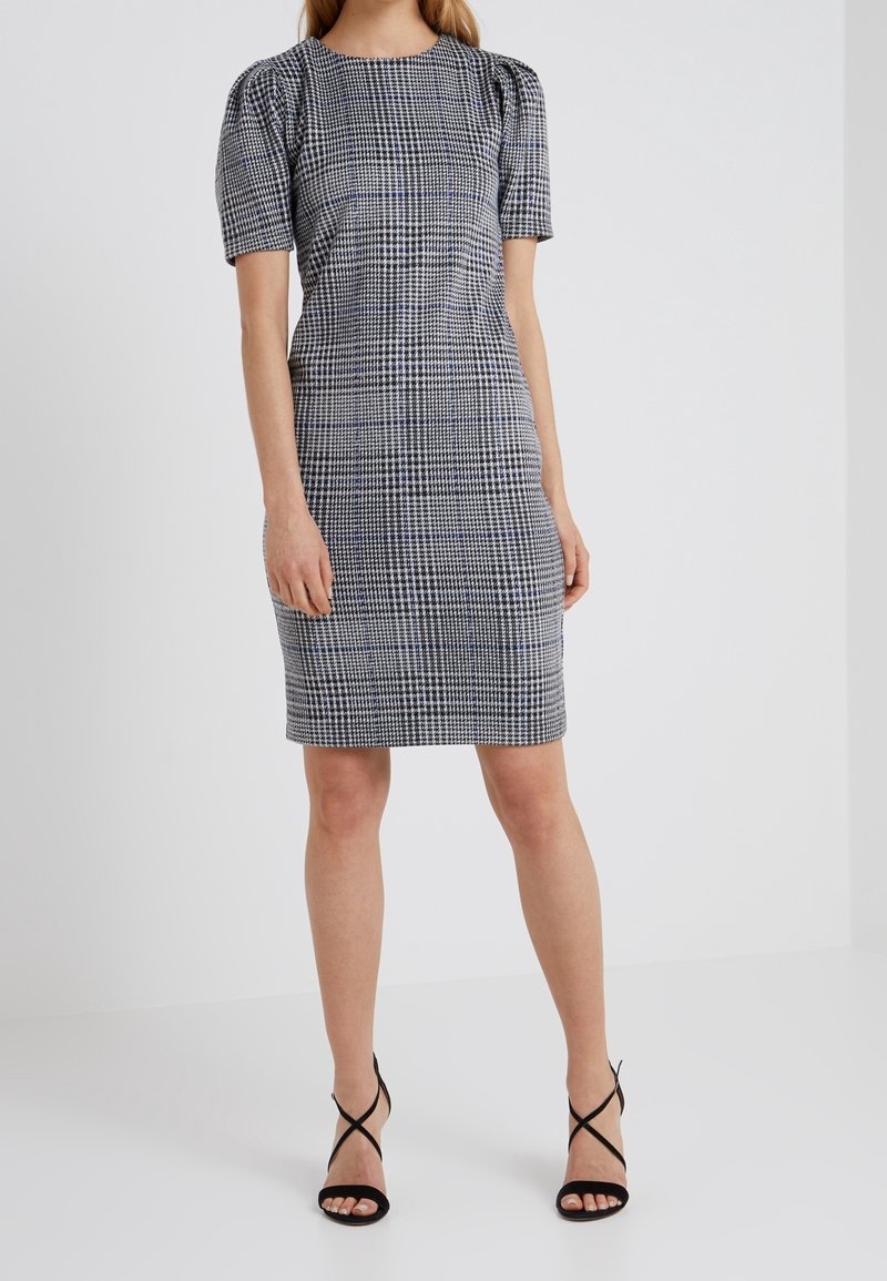 Femme portant une robe à carreaux noir et blanc longueur genou avec des manches courtes bouffantes et des escarpins noirs à brides.