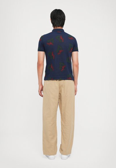 Ναυτικό πόλο κοντομάνικο μπλουζάκι με αφηρημένα κόκκινα και πράσινα μοτίβα, συνδυασμένο με μπεζ παντελόνι σε φαρδιά γραμμή και λευκά παπούτσια. Προβολή από πίσω.