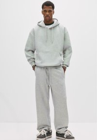 Mand står iført en lysegrå hættetrøje og matchende sweatpants, sorte sneakers, hænderne i lommerne, mod en ensfarvet hvid baggrund.