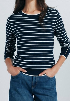 Woman wearing navy blue and white horizontal striped long-sleeve sweater with gold buttons on cuffs and blue denim jeans, hands in pockets.