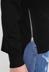 Zwarte gebreide sweater met een zijrits met zilveren beslag, geribbelde boorden en onderkant, en een relaxte pasvorm. Gecombineerd met versleten blauwe jeans.
