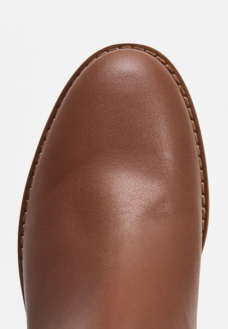 Primo piano di una scarpa in pelle marrone liscia con cuciture visibili lungo il bordo della suola su uno sfondo bianco.