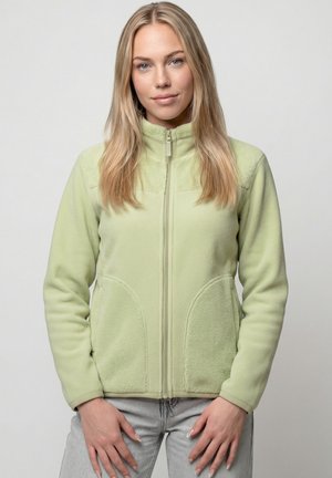 Blonde Frau trägt eine hellgrüne Fleecejacke mit Reißverschluss und hellgraue Jeans, steht mit entspannten Händen vor dem Körper.