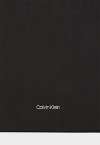 Černá textilní taška s texturovaným povrchem a stříbrným logem "Calvin Klein" umístěným na prominentním místě.