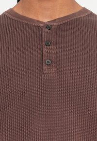 Nahaufnahme einer Person, die ein mauvefarbenes Waffelstrick-Henley-Shirt mit drei schwarzen Knöpfen am Vorderfalz trägt.