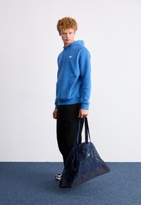 Sweatshirt azul com bolso frontal e logotipo, combinado com calças pretas e uma bolsa de rede azul marinho com painéis texturizados e duas alças.
