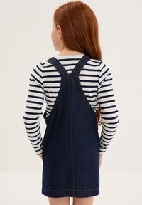 Abito salopette in denim di tessuto blu scuro, con spalline regolabili e una vestibilità attillata, indossato sopra una camicia a maniche lunghe bianca e blu navy a righe.