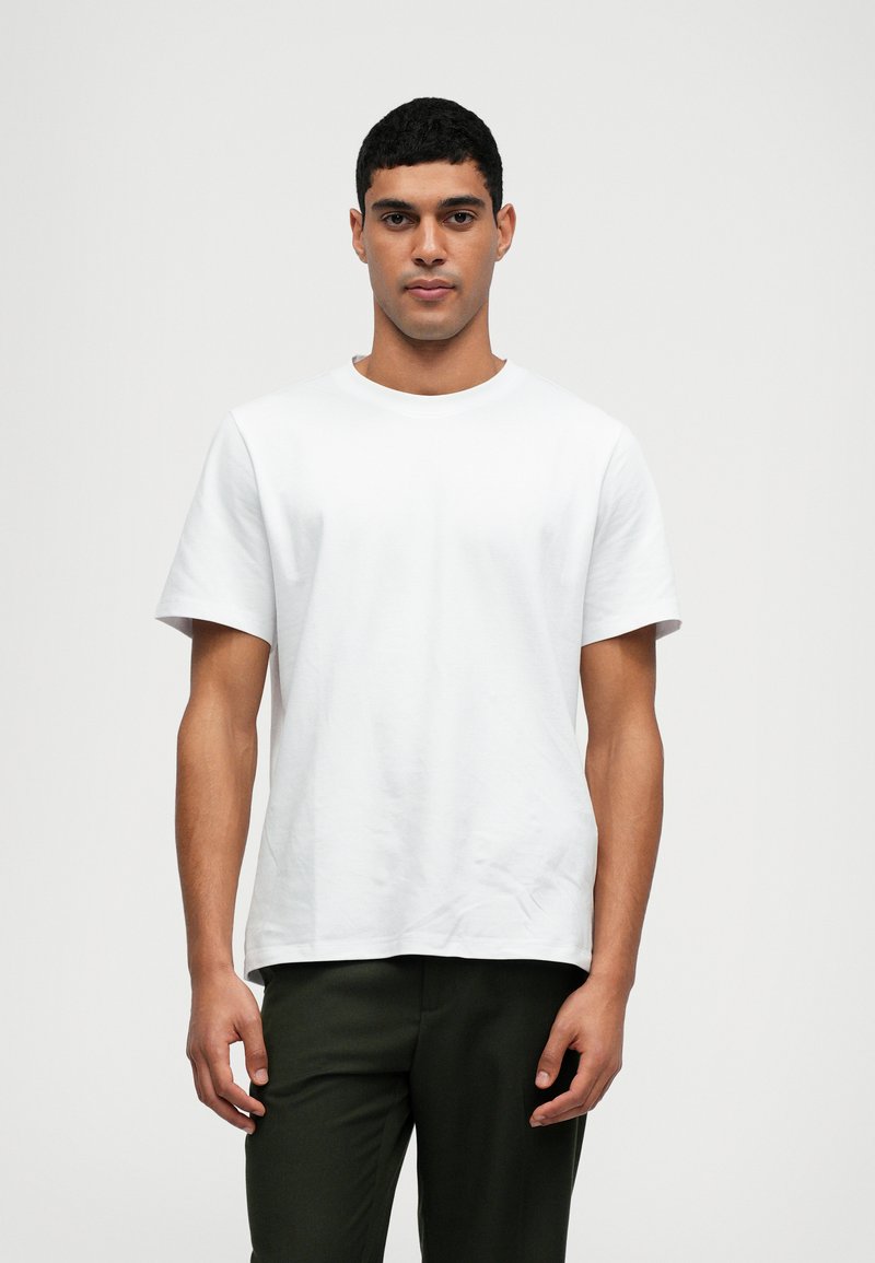 Λευκό βαμβακερό T-shirt με στρογγυλό λαιμό, κοντά μανίκια και άνετη εφαρμογή, συνδυασμένο με σκούρους πράσινους παντελόνες. Λείος υφή, μινιμαλιστικός σχεδιασμός.