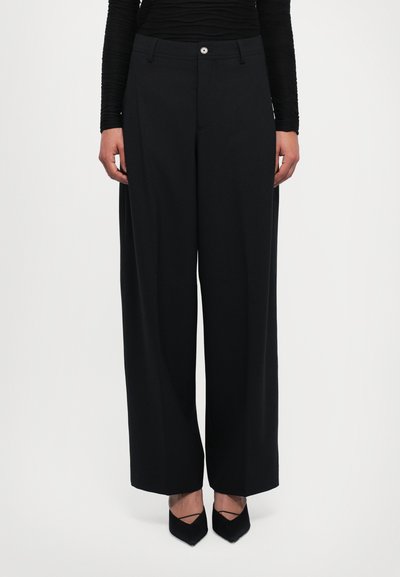 Woman wearing black wide-leg trousers, long-sleeve black top, and black pointed-toe heels standing against white background.