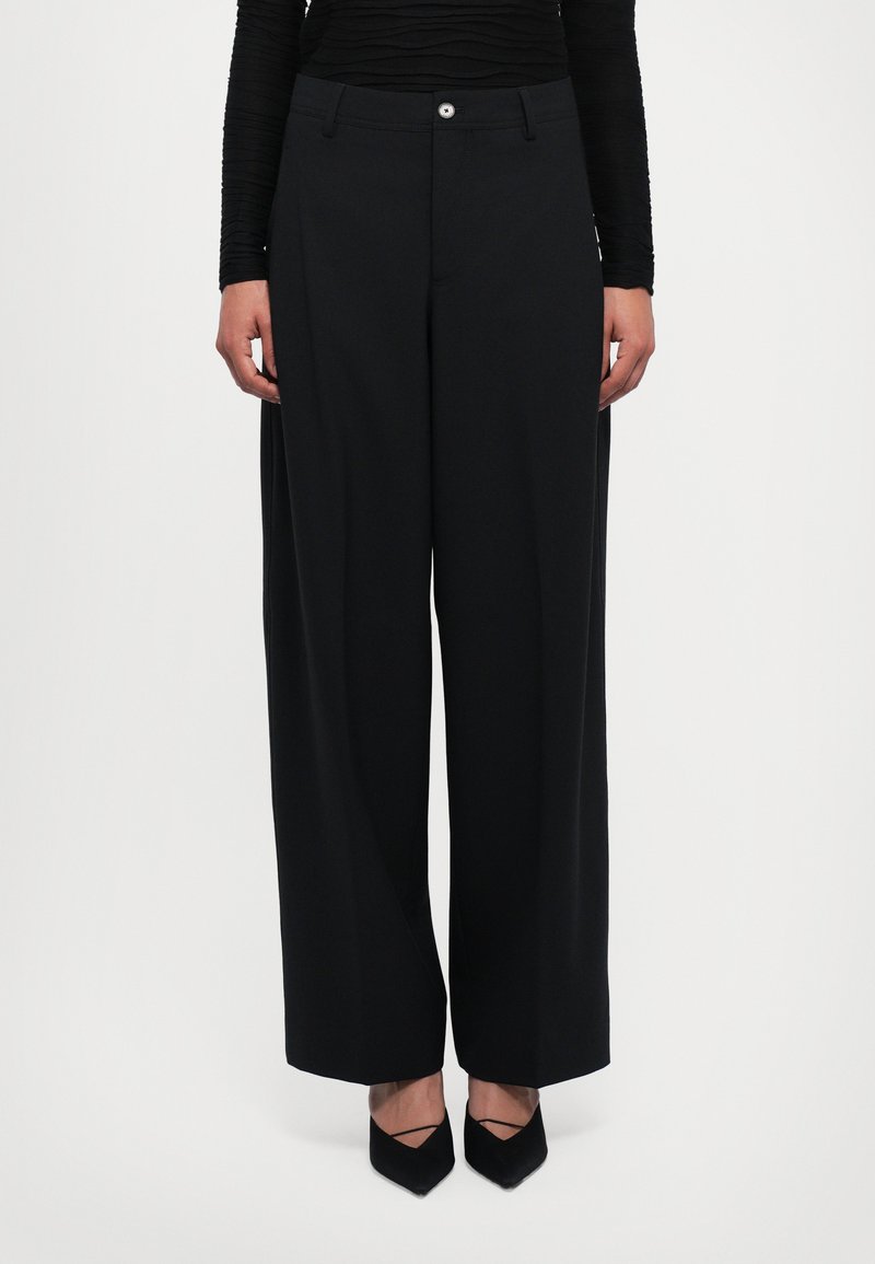 Woman wearing black wide-leg trousers, long-sleeve black top, and black pointed-toe heels standing against white background.