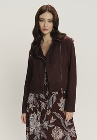 Giacca cropped in finta suede borgogna con chiusura a zip e colletto a punta, abbinata a un vestito con stampa floreale multicolore.