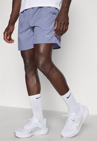 Pantalones cortos de running de Nike en púrpura claro, con un logo de swoosh gris. Combinados con calcetines deportivos blancos y zapatillas deportivas blancas.
