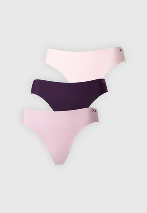 Femme aux longs cheveux foncés, debout pieds nus, portant un soutien-gorge mauve et des sous-vêtements assortis sur un fond blanc uni.
