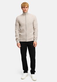 Blond mężczyzna ubrany w beżowy sweter z zamkiem błyskawicznym i prążkowanym wzorem, czarne spodnie i białe trampki, stojący na tle jednolitego białego tła.