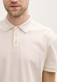 Hellbeige Polohemd aus strukturiertem Stoff, mit knöpfbarem Hals und kontrastierendem grün-weiß gestreiften Kragen mit Logodetail.
