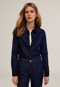 Camisa de mezclilla azul oscuro con botones en la parte delantera, cuello adornado y puños con tachuelas decorativas. Silueta ajustada, textura suave.