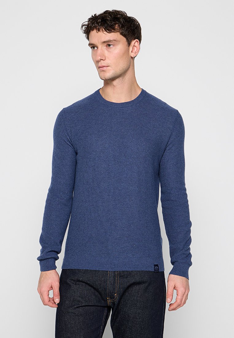 Uomo che indossa un maglione blu a maniche lunghe aderente con collo rotondo e jeans in denim scuro, in piedi contro uno sfondo chiaro e semplice.