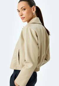 Veste cropped en simili cuir beige avec un col à revers, une poche latérale et un ourlet élastique. Texture lisse et lignes modernes épurées.