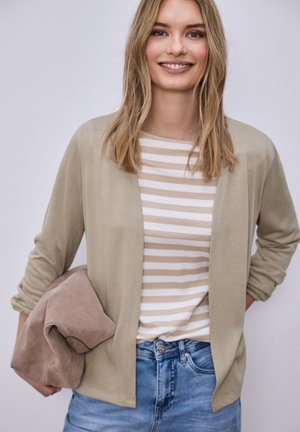 Donna sorridente con capelli biondi che indossa un cardigan beige, una maglia a righe beige e bianca, jeans blu, che tiene una giacca beige piegata sul braccio.