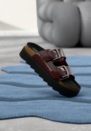 Sandale en cuir marron avec doubles boucles argentées et semelle noire épaisse, posée sur un tapis bleu texturé dans un environnement intérieur moderne.